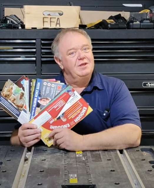 A man holding a large stack of glossy campaign mailers in front of shelves full of more mail.